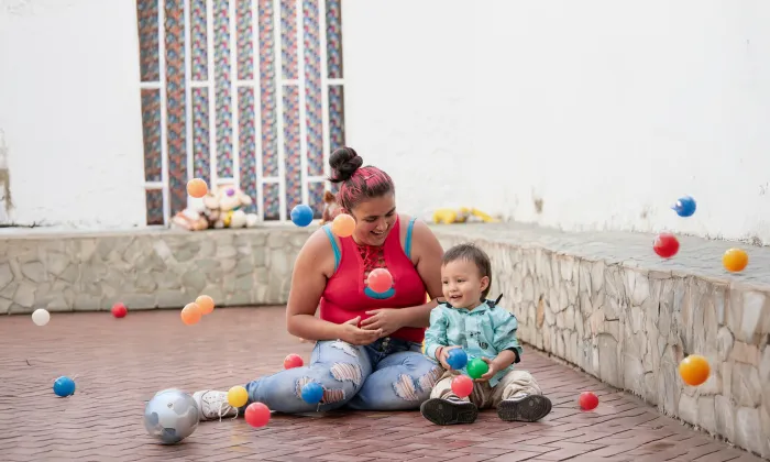 Mujer sentada en el suelo jugando con su hijo pequeño, rodeados de pelotas de colores en un patio con paredes blancas.