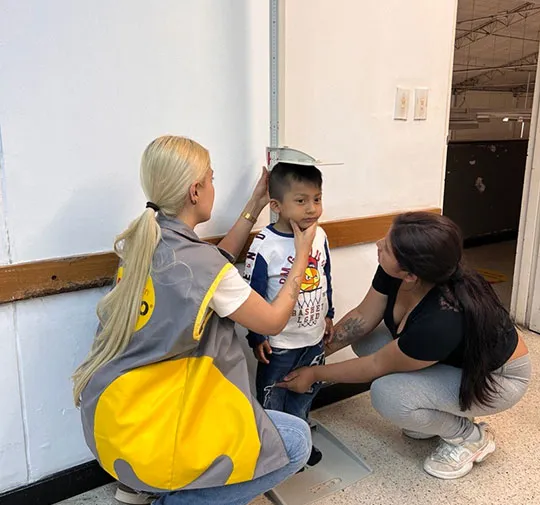 Una voluntaria con chaleco amarillo mide la estatura de un niño que está de pie sobre una báscula, mientras la madre del niño se agacha a su lado en un espacio interior.