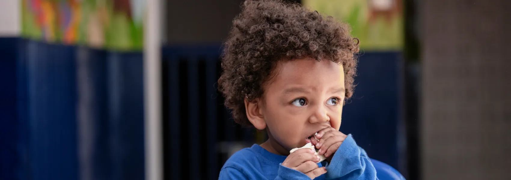 Niño pequeño con cabello rizado vistiendo una camiseta azul, comiendo un bocadillo mientras observa con curiosidad hacia un lado, con un fondo desenfocado.